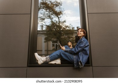 Cheerful pretty young caucasian student sits on windowsill in city, holds phone and coffee. Beauty is dressed denim jacket, jeans and white shoes. Fashion, technology and people concept. - Powered by Shutterstock