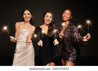 Cheerful pretty young girlfriends multiracial women wearing elegant dresses have New Year 2024 party celebration on black background, holding bengal lights and smiling at camera - Powered by Shutterstock