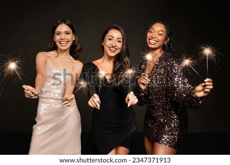 Cheerful pretty young girlfriends multiracial women wearing elegant dresses have New Year 2024 party celebration on black background, holding bengal lights and smiling at camera