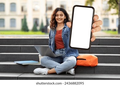 Cheerful pretty young indian woman student sitting on stairs with laptop on her lap showing big phone with blank white screen, university campus. Educational mobile app, online course, e-education - Powered by Shutterstock