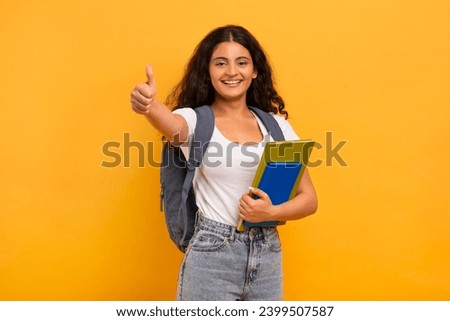 Cheerful pretty young indian woman student with backpack and books in her hand showing thumb up and smiling at camera, posing isolated on yellow background. Education, school, college