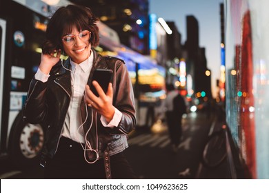 Cheerful pretty young woman in cool eyeglasses and trendy wear walking on metropolis street with night lights enjoying online playlist songs in earphones and reading sms with good news on smartphone