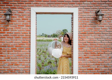 Cheerful Pretty Young Woman With Hair Tied By Handkerchief Standing And Holding Carry Box Of Her Lovely Prairie Dog Pet In Front Of Vintage Red Brick Door In Beautiful Flower Garden With Blue Sky.