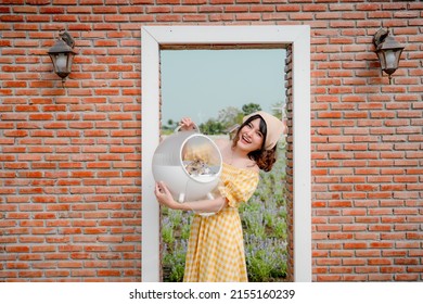 Cheerful Pretty Young Woman With Hair Tied By Handkerchief Standing And Holding Carry Box Of Her Lovely Prairie Dog Pet In Front Of Vintage Red Brick Door In Beautiful Flower Garden With Blue Sky.