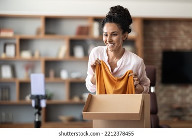 Cheerful Pretty Young Woman Unpacking Parcel And Recording Video, Streaming Via Cell Phone Set On Tripod, Excited Lady Having Video Chat With Friend, Showing Her New Clothes And Smiling, Copy Space