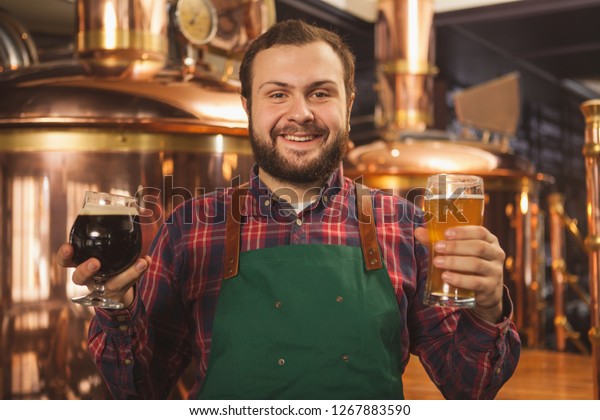 Cheerful professional brewer in an apron smiling\
happily to the camera holding two glasses of beer, enjoying working\
at his microbrewery. Happy bartender serving two beers. Friendly\
service concept