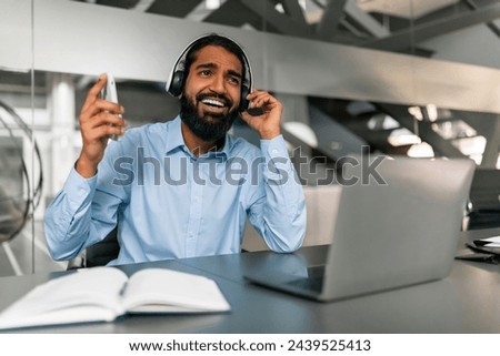 Cheerful professional using a headset and holding a smartphone in a modern office setting, indicating remote communication