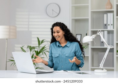 Cheerful professional woman in a blue shirt engaging in a video call on her laptop in a well-lit modern office setting. - Powered by Shutterstock