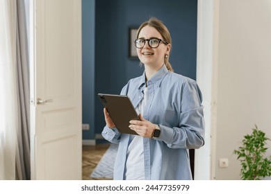 A cheerful professional woman with a tablet, smartwatch, and eyeglasses, wearing a light blue shirt, stands in an airy room with a backdrop of tasteful decor - Powered by Shutterstock