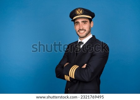Cheerful and professional young male pilot in a studio setting, wearing a pilot uniform, smiling confidently against a vibrant blue background, showcasing the spirit and enthusiasm of aviation