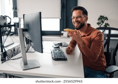 Cheerful programmer multitasks by recording a voice message on his smartphone while working in a modern office. The image captures a moment of innovation and productivity in a corporate setting
