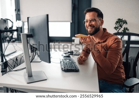 Cheerful programmer multitasks by recording a voice message on his smartphone while working in a modern office. The image captures a moment of innovation and productivity in a corporate setting