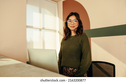 Cheerful receptionist standing at the front desk of a co-working space. Friendly female assistant looking at the camera while working at the reception in a modern office.