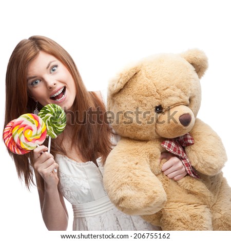 cheerful red-haired woman in short white dress holding colored lollipops and big soft toy bear isolated on white