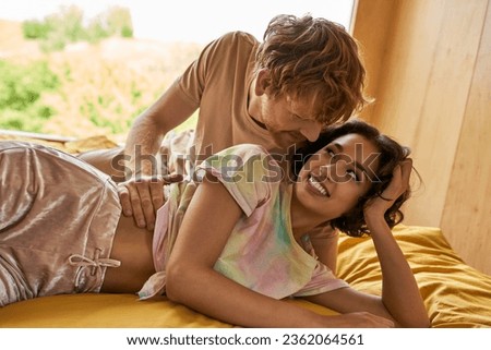 cheerful redhead man flirting with asian girlfriend and lying on bed together, having a good time