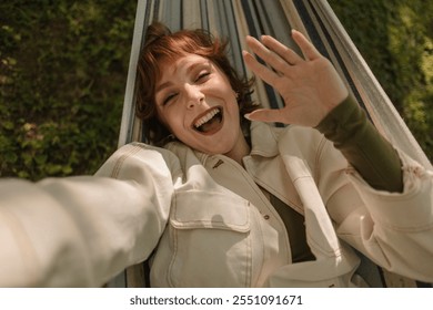 Cheerful redhead woman with freckles is lying in a hammock, taking a selfie and waving her hand, enjoying a sunny day outdoors - Powered by Shutterstock