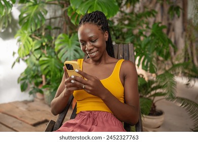 Cheerful relaxed African American girl rest takes break with smartphone among grows indoor plants spending time online on Internet. Happy Black woman smiling using cellphone text social media message. - Powered by Shutterstock