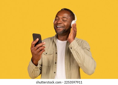 Cheerful relaxed mature african american man in casual and wireless headphones looking at phone, enjoy spare time, listen favorite music, isolated on yellow background, studio. Audio app, ad and offer - Shutterstock ID 2253597911