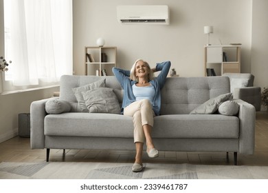 Cheerful relaxed retired old woman resting on grey soft couch under domestic conditioner in stylish cozy apartment interior, breathing cool fresh air, cold comfortable temperature in living room - Powered by Shutterstock