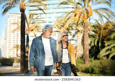 Cheerful retired couple enjoying a leisurely stroll through a vibrant city park adorned with palm trees, savoring the pleasant weather and each other's company during their golden years