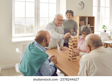 Cheerful retired friends playing jenga board game at home. Happy senior men and women sitting around table playing and communicating in living room. Elderly people leisure activities, nursing home - Powered by Shutterstock