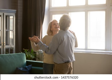 Cheerful retired spouses husband and wife laughing dancing in living room, happy romantic old middle aged couple enjoying slow dance having fun celebrating anniversary or new house purchase at home - Powered by Shutterstock