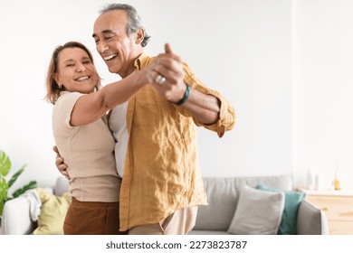 Cheerful retired spouses husband and wife dancing and laughing in living room, happy romantic couple enjoying slow dance, celebrating anniversary at home - Powered by Shutterstock