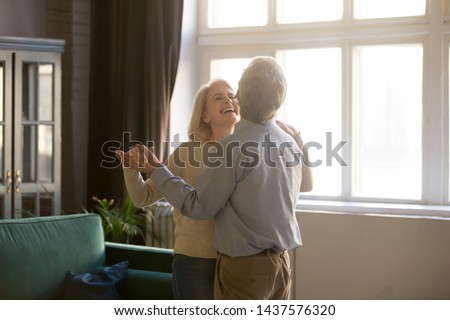 Cheerful retired spouses husband and wife laughing dancing in living room, happy romantic old middle aged couple enjoying slow dance having fun celebrating anniversary or new house purchase at home