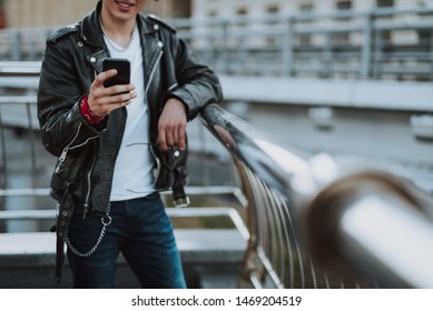 Cheerful Rocker In Leather Jacket Standing Near The Banister And Using Smartphone