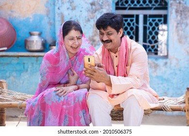Cheerful rural indian couple using smart phone sit on bed. farmer husband wife watching at mobile screen, man woman smiling. Internet video call, village people use cellphone Technology. digital india - Powered by Shutterstock
