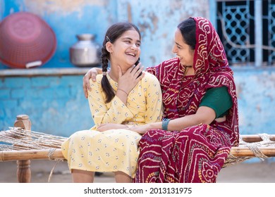 Cheerful rural Indian mother and young child daughter laughing having a good time together, sitting on traditional bed, Cute adorable girl kid in braided hair and mum in red sari, happy parent bonding