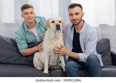 Cheerful Same Sex Couple Sitting On Couch With Dog