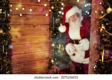 Cheerful Santa Claus looks into a wooden house, beautifully decorated for Christmas, with a big bag of presents. 