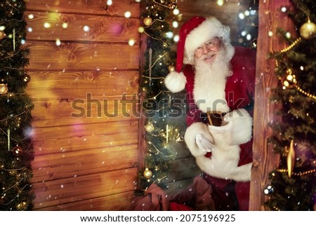 Cheerful Santa Claus looks into a wooden house, beautifully decorated for Christmas, with a big bag of presents. 