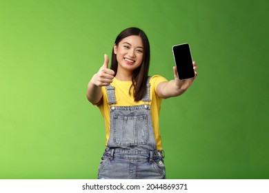 Cheerful Satisfied Cute Asian Brunette Girl Pleased Awesome Smartphone Game, Show Telephone Display, Give Thumb-up Like Sign, Smiling Broadly, Introduce Awesome Online Application, Green Background