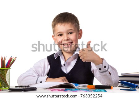 Cheerful satisfied pupil sitting at the desk with thumb up surrounded with stationery