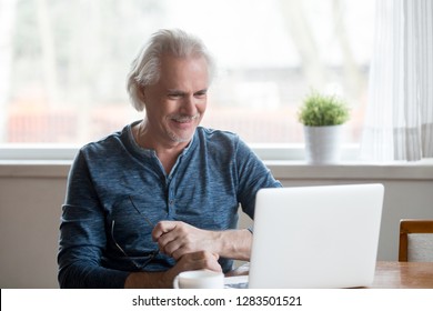 Cheerful Satisfied Retired Mature Person Sitting At Table In The Kitchen Working On Computer Received Good Positive News From Bank Using Laptop For Entertaining Watching Online Program Or Funny Video