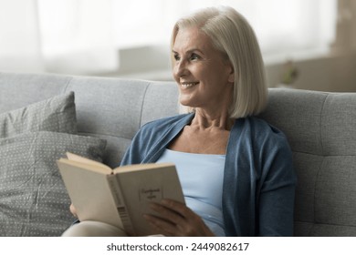 Cheerful senior 60s woman sit on cozy sofa read interesting book, looking away, enjoy literature hobby, daydreaming, ponder over plot of novel, spend leisure time alone at home. Pastime, book-lover
