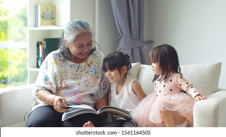 Cheerful Senior Asian Grandma Sitting On Couch And Reading A Book With Two Her Granddaughter With Fun In The Living Room At Home 