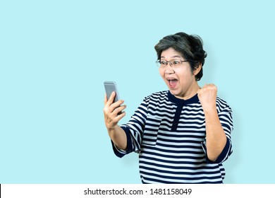 Cheerful Senior Asian Woman Holding And Looking Smartphone On Isolated Background Feeling Win ,celebrating And Victory. Older Female Lifestyle Concept  Blue Background.