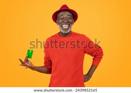 A cheerful senior black man with a bright white smile, wearing a red hat and sweater, holds a green credit card, standing against an orange background, studio. Finance, money