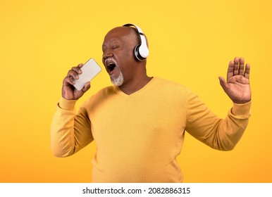 Cheerful Senior Black Man Listening To Music In Wireless Headphones, Singing, Using Smartphone As Microphone, Having Fun On Orange Studio Background, Mockup For New Mobile App