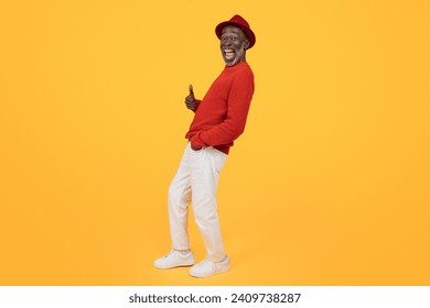 Cheerful senior Black man wearing a red sweater and matching hat, giving a thumbs up and looking to the side with a wide smile on a plain yellow background, full length - Powered by Shutterstock