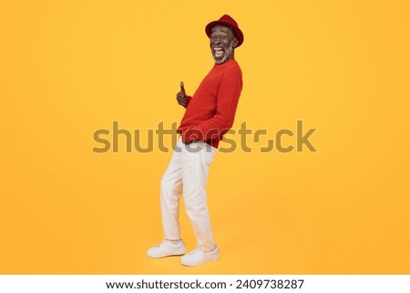 Cheerful senior Black man wearing a red sweater and matching hat, giving a thumbs up and looking to the side with a wide smile on a plain yellow background, full length