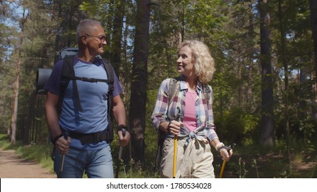 Cheerful senior caucasian couple hiking in park with backpacks, enjoying adventure. Mature man and woman nordic walking in forest. Active lifestyle, health, retirement concept