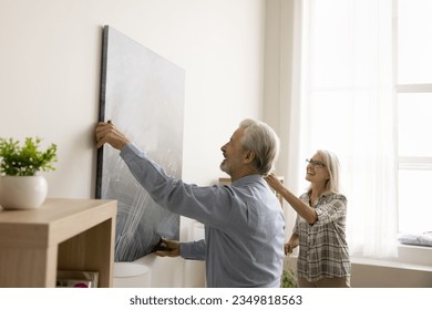 Cheerful senior couple decorating apartment, hanging large picture on white wall, laughing, having fun, working on home interior contemporary design, enjoying domestic activities in new house - Powered by Shutterstock