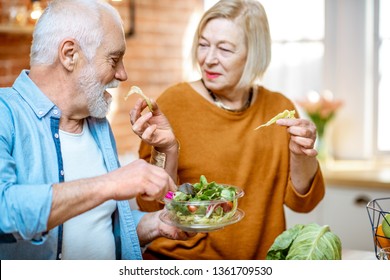 Cheerful Senior Couple Eating Salad Standing Together With Healthy Food On The Kitchen At Home. Concept Of Healthy Nutrition In Older Age