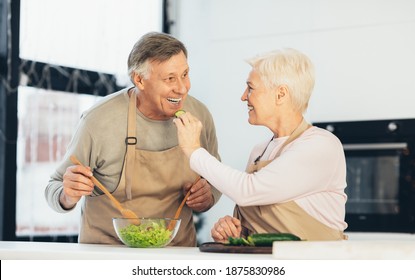Cheerful Senior Couple Feeding Each Other And Laughing Enjoying Cooking Together In Modern Kitchen At Home. Older Spouses Having Fun Tasting Food. Family Dinner Preparation