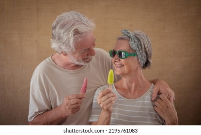 Cheerful Senior Couple Holding A Colored Popsicle Stick Looking In The Eyes, Happy Retirees Enjoying Retirement Eating A Fresh Popsicle