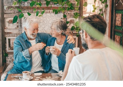 Cheerful senior couple and mature son inside a cozy cafeteria having a break with coffee, cappuccino and fruit cake. Three people enjoying stay together - Powered by Shutterstock